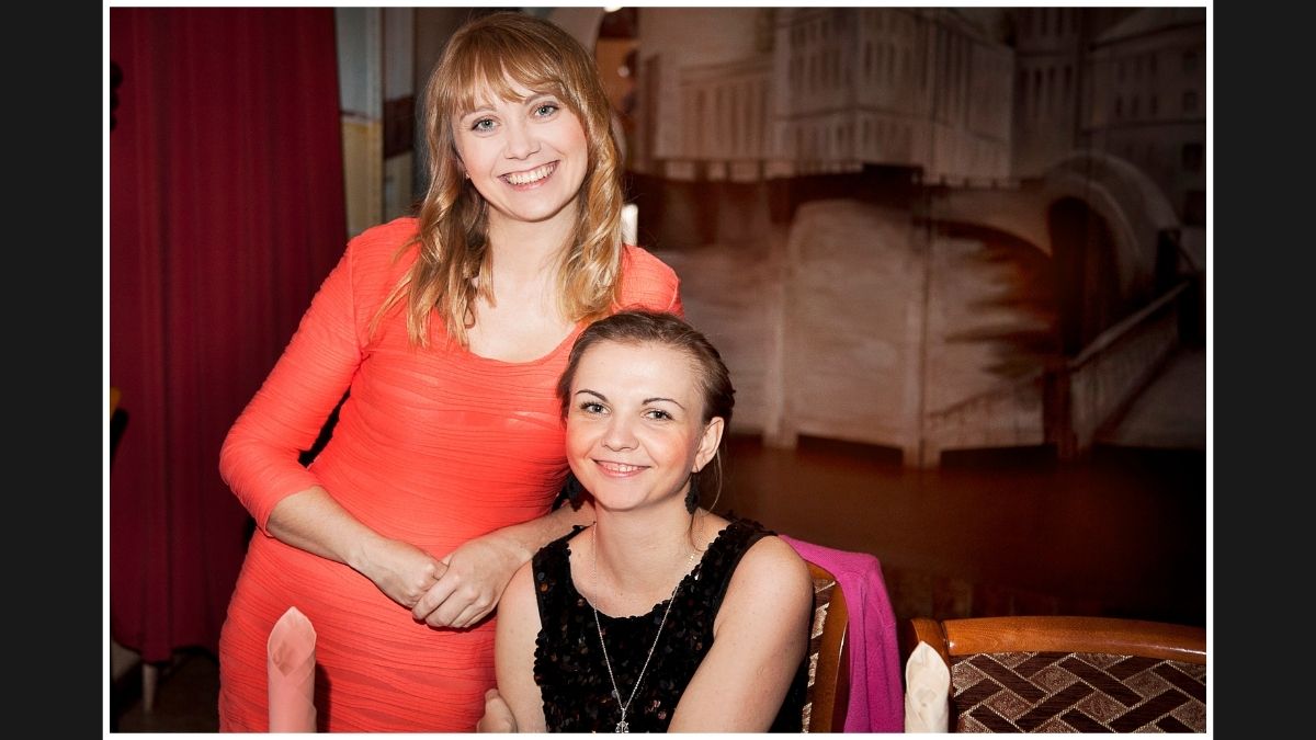 Two women smiling together, with one standing and the other sitting, in a warmly lit environment.