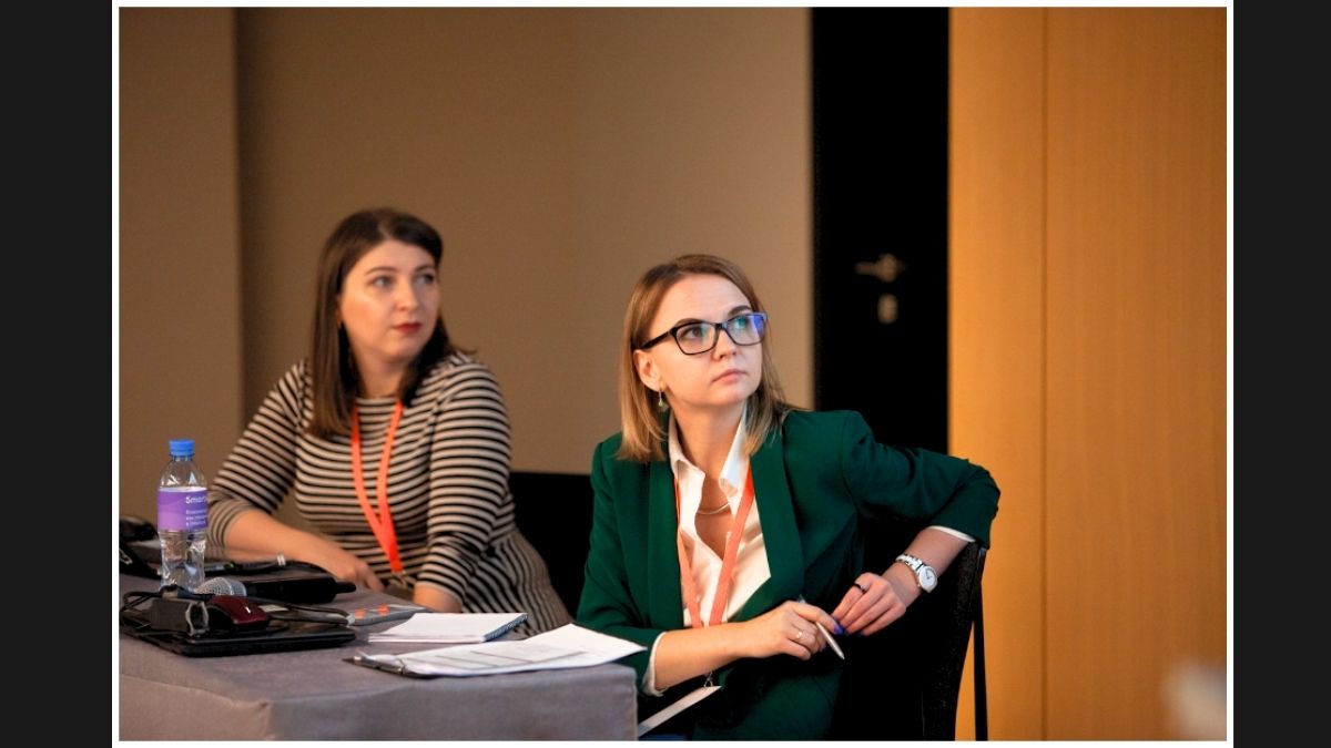 Two focused women at a conference, listening intently, with notebooks and pens ready.