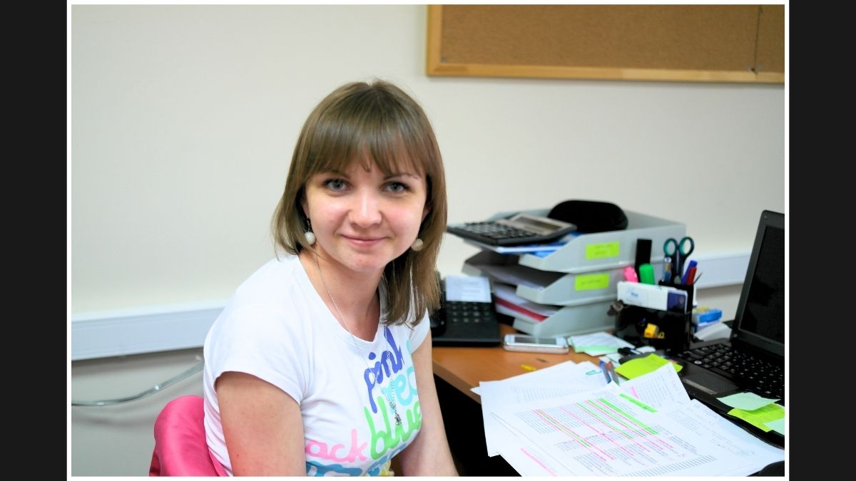 Woman in a casual white T-shirt, seated at a desk, looking at the camera with a gentle smile.