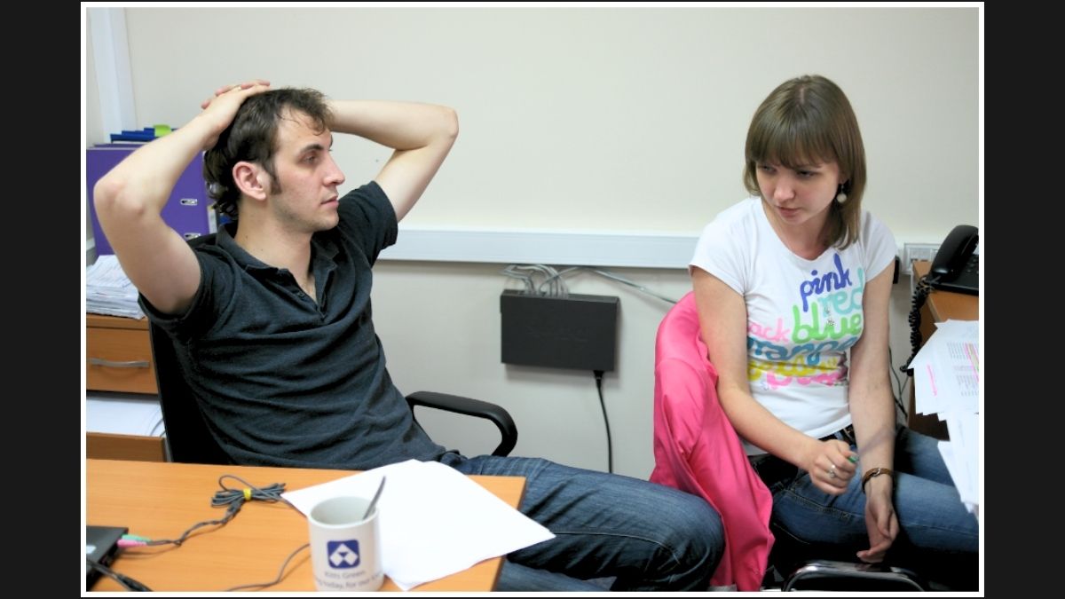 Two colleagues in a casual office discussion; one looks thoughtful with hands on his head, while the other listens.