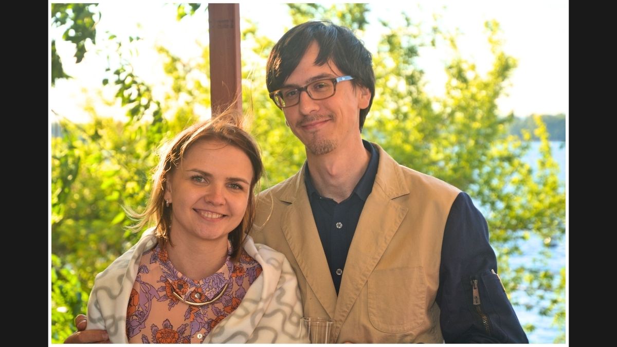 A smiling couple posing outdoors with lush greenery behind them, enjoying a sunny day.